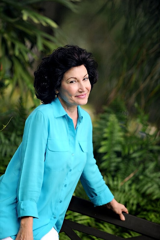 Smiling woman in turquoise shirt surrounded by greenery.