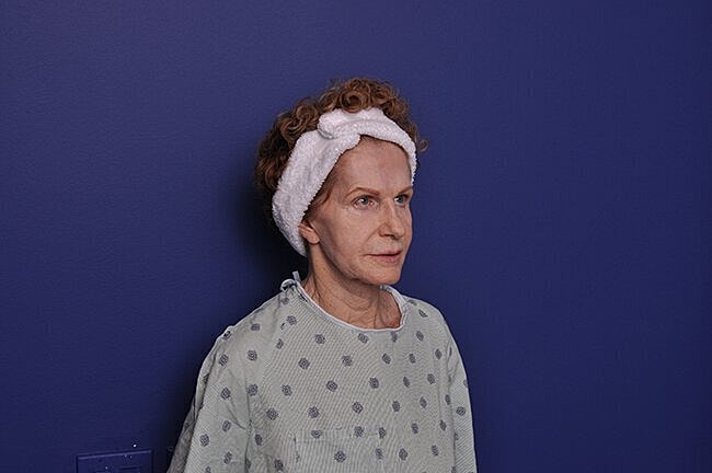 Older woman in hospital gown against purple wall.