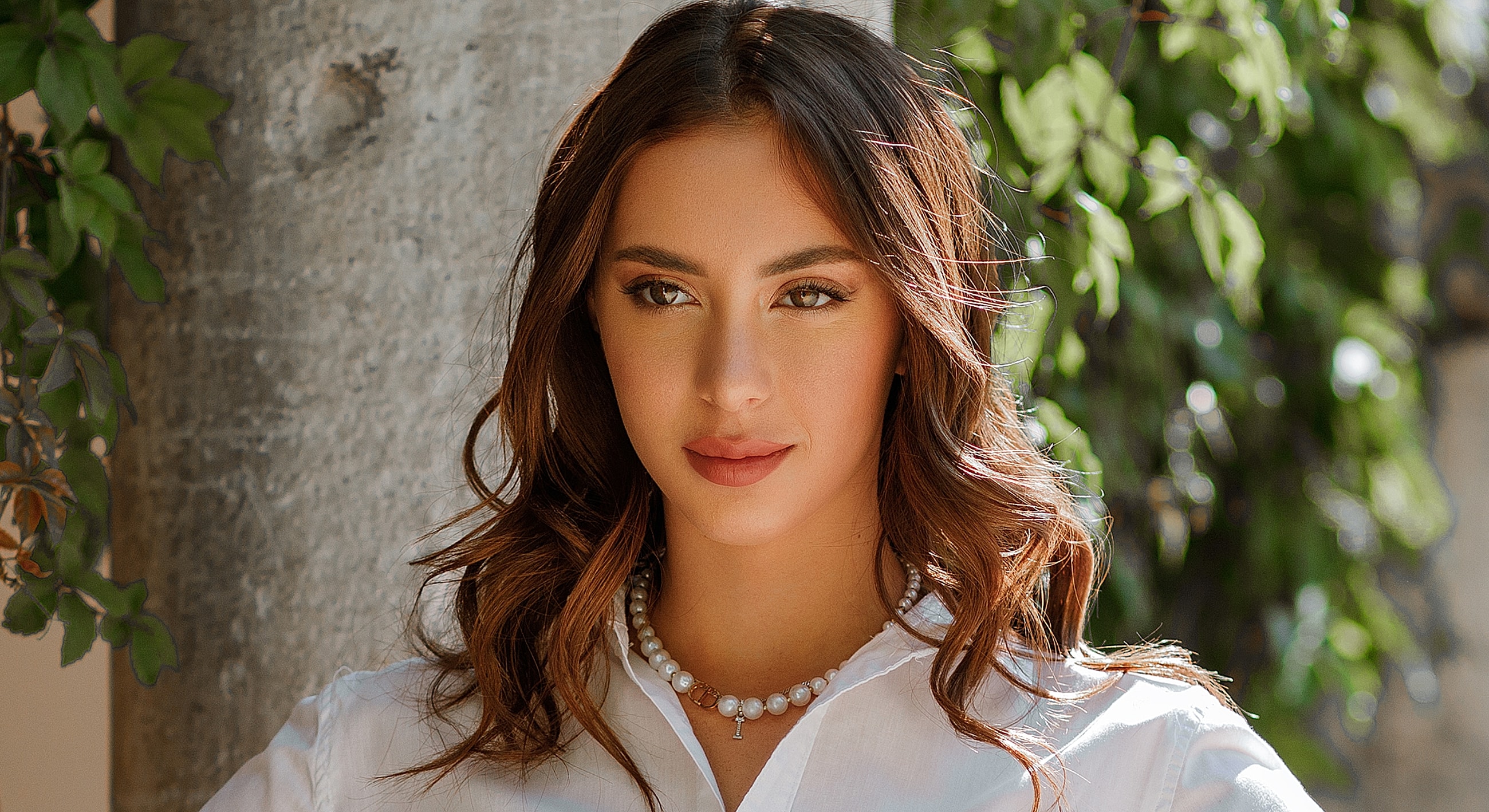 Woman with wavy hair posing outdoors by tree.